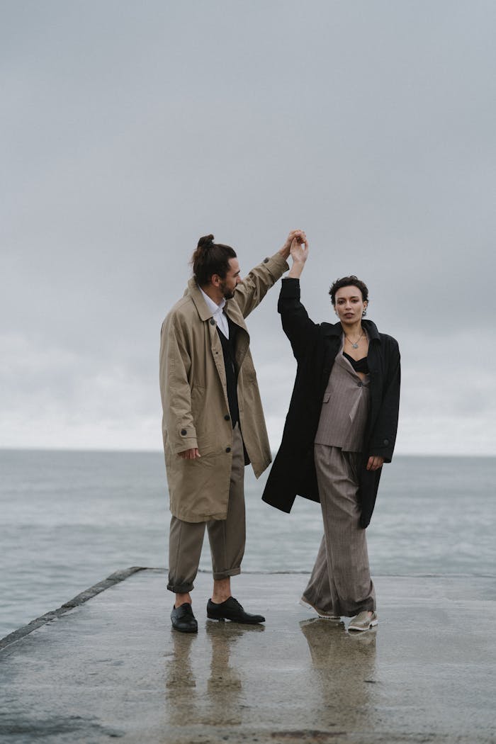 An elegant couple dances on a pier by the sea in trench coats, exuding romance and style.